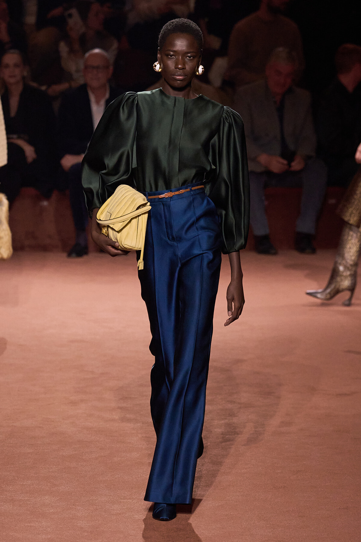 Model walking a runway in a dark green puff-sleeve blouse, cobalt satin pants, tan belt, and a pale yellow clutch with gold earrings nearby.