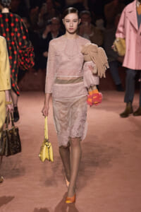 Runway model in pale pink lace dress carries a stuffed doll and yellow handbag while walking the catwalk.
