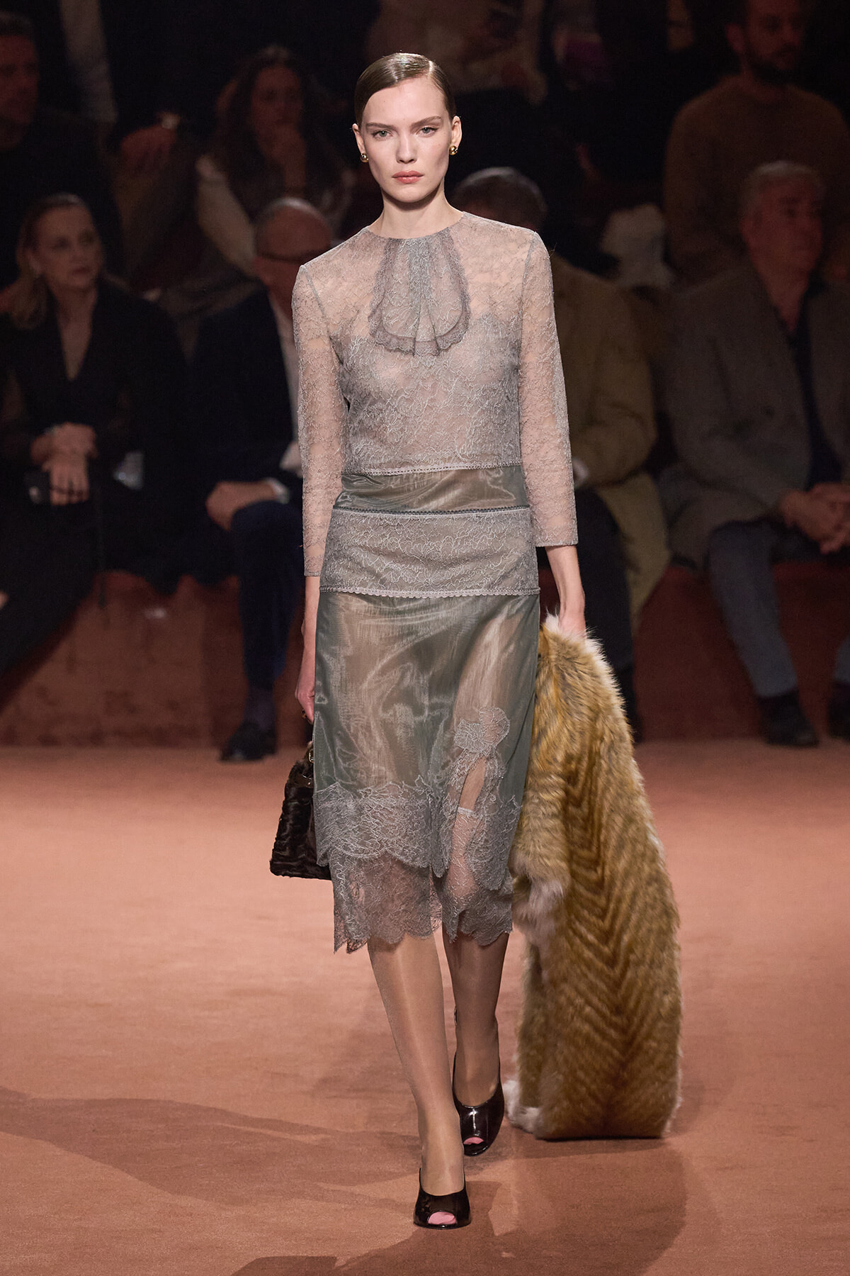 Model walks the runway in a pale gray lace dress with sheer long sleeves, a ruffled high neckline, and a scalloped hem, carrying a dark handbag.