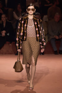 Model on runway wearing a beige polka-dot midi dress with a fur coat featuring pink pom-poms, oversized sunglasses, and a brown handbag.