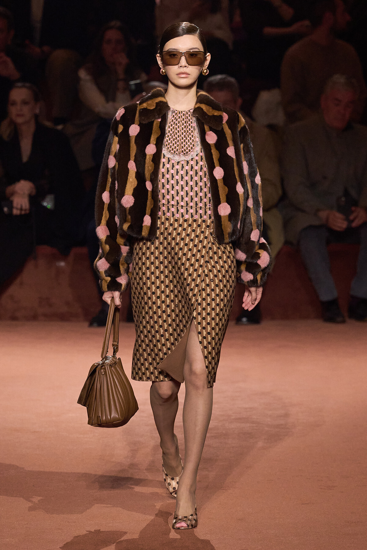 Model on runway wearing a beige polka-dot midi dress with a fur coat featuring pink pom-poms, oversized sunglasses, and a brown handbag.