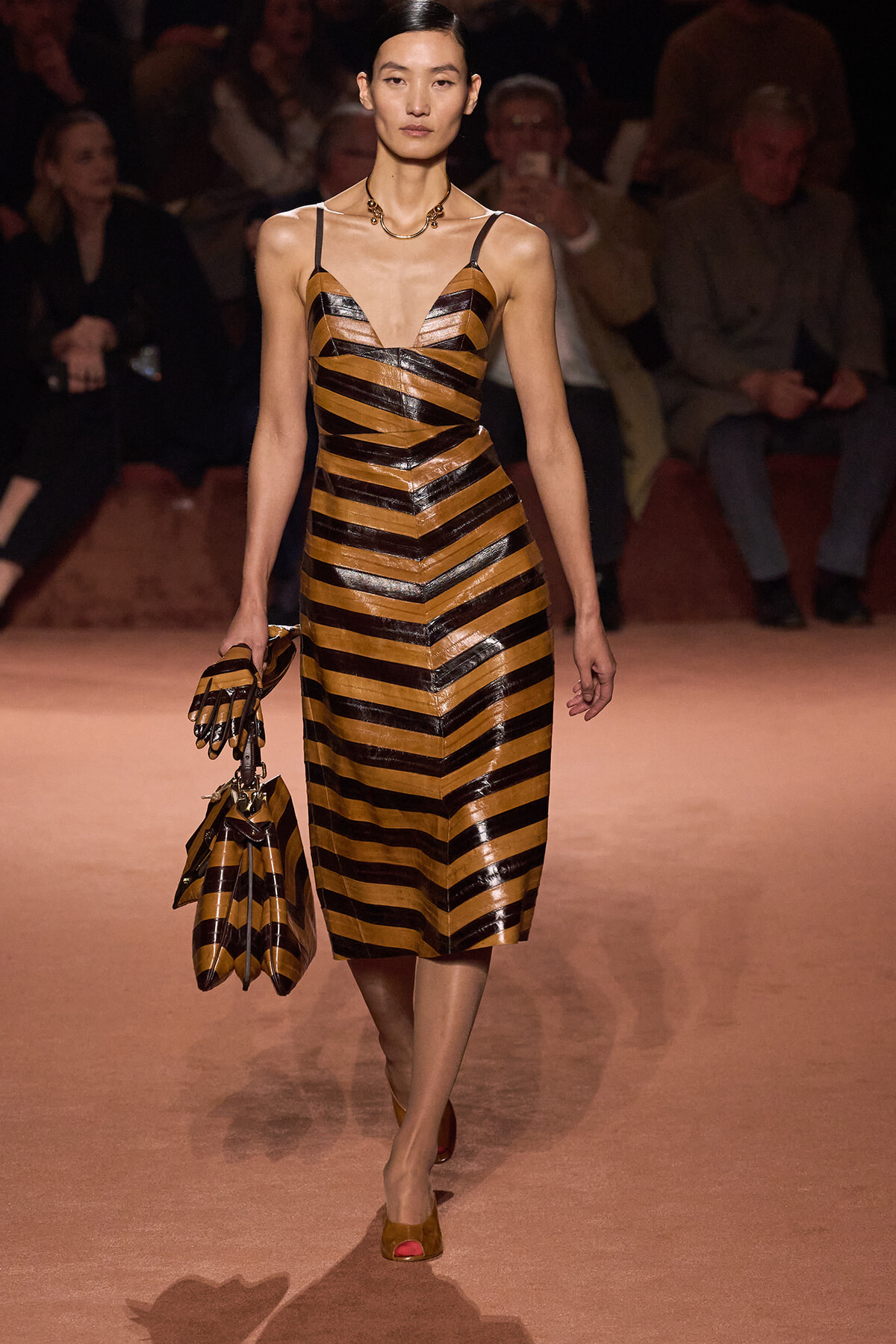 Model walking a fashion runway in a gold and black striped midi dress with thin straps and matching gloves, carrying a striped handbag group.”]} JSON error: I provided invalid JSON. Let's correct. {