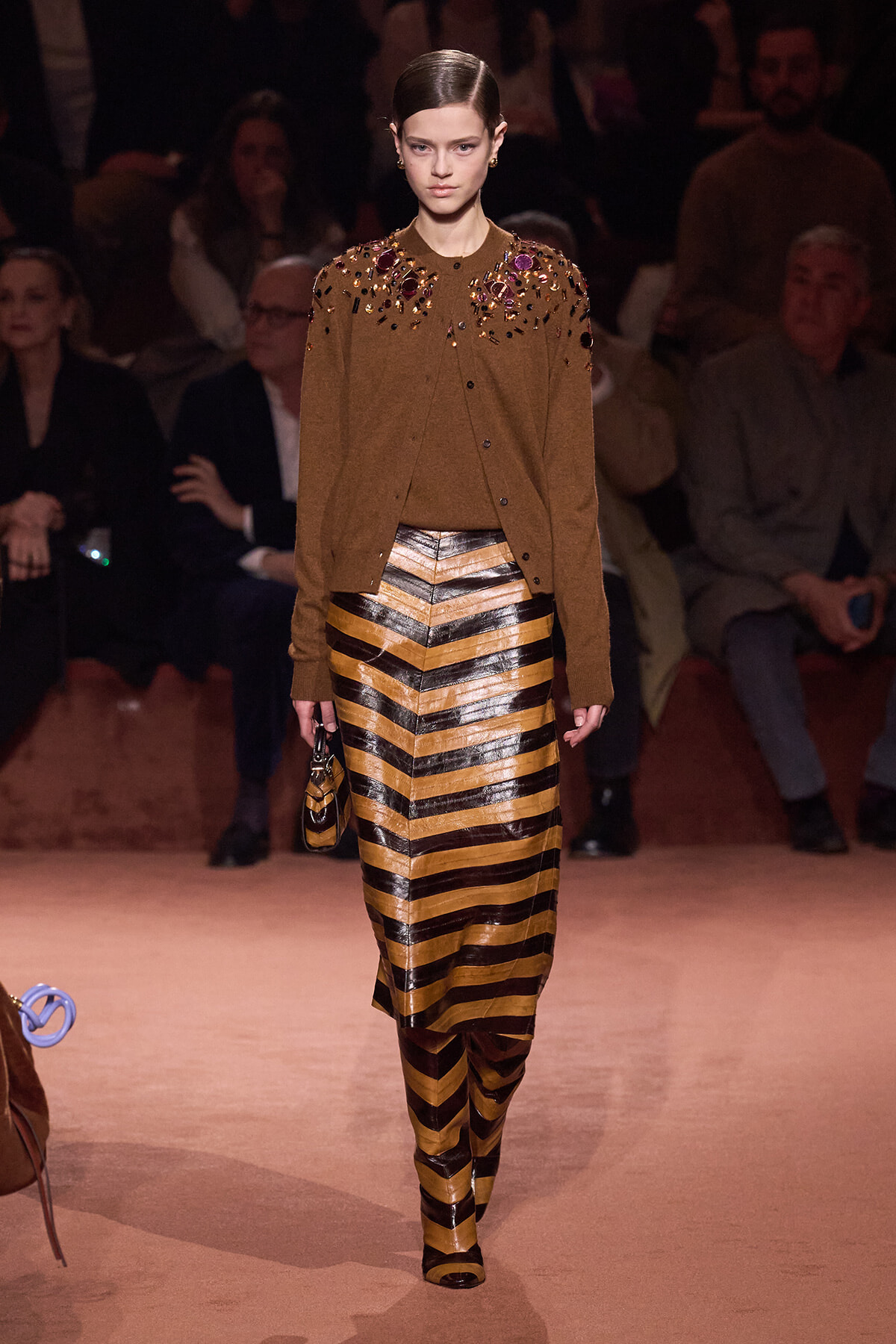 Model walking a fashion runway in a brown knit cardigan adorned with colorful jewels, paired with a gold and black chevron striped skirt and matching tights.