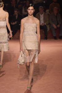 Model walking a runway in a beige sheer lace dress with sculpted panels, holding a matching bag on her left side.