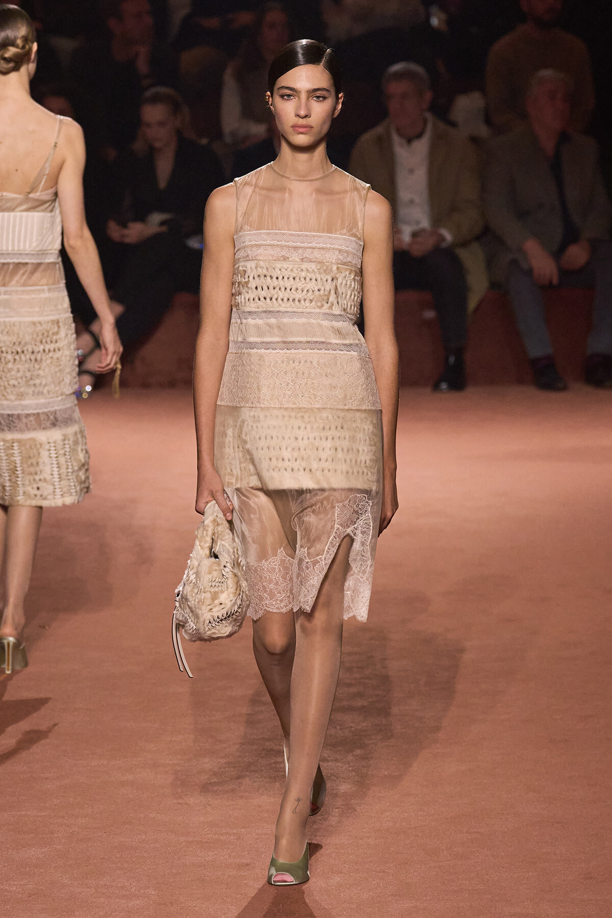 Model walking a runway in a beige sheer lace dress with sculpted panels, holding a matching bag on her left side.