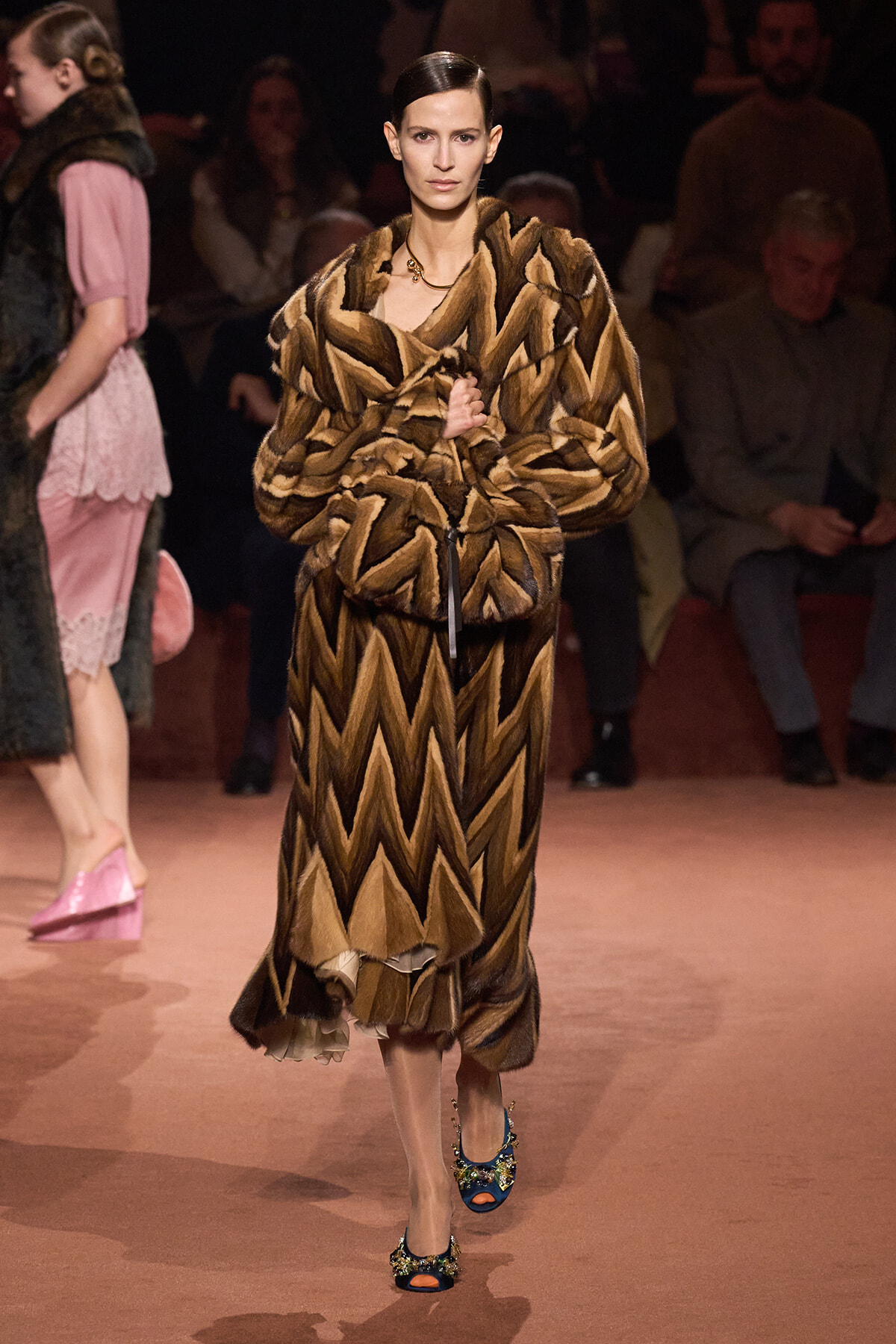 Model on a runway wearing a bold chevron-pattern brown fur coat and blue embellished heels, audience in the background.