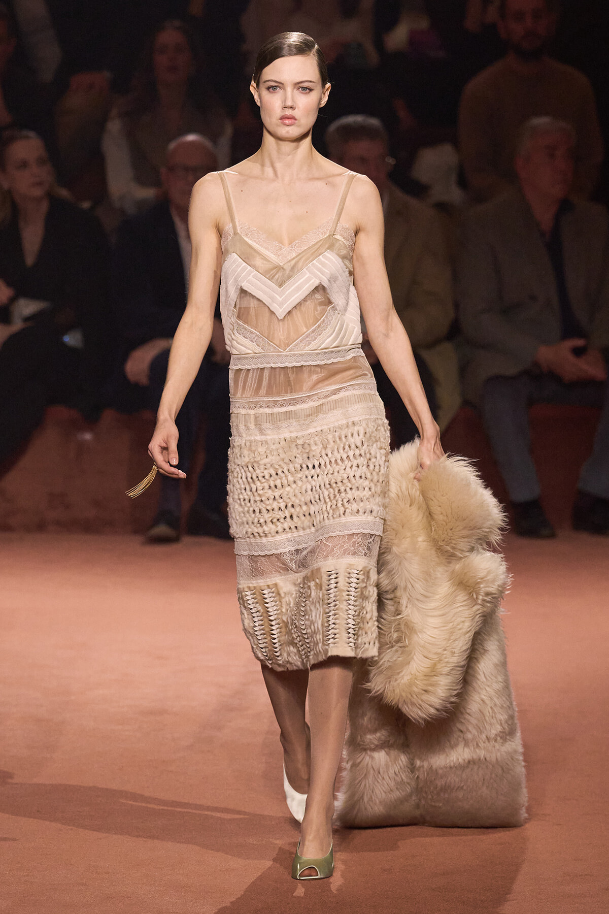 Model walking a runway in a beige lace cocktail dress with sheer panels, holding a large cream fur coat.