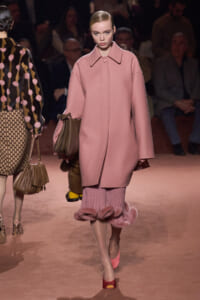 Model walks a runway in a oversized pink coat, carrying a brown handbag, with pink ruffled hem visible beneath.