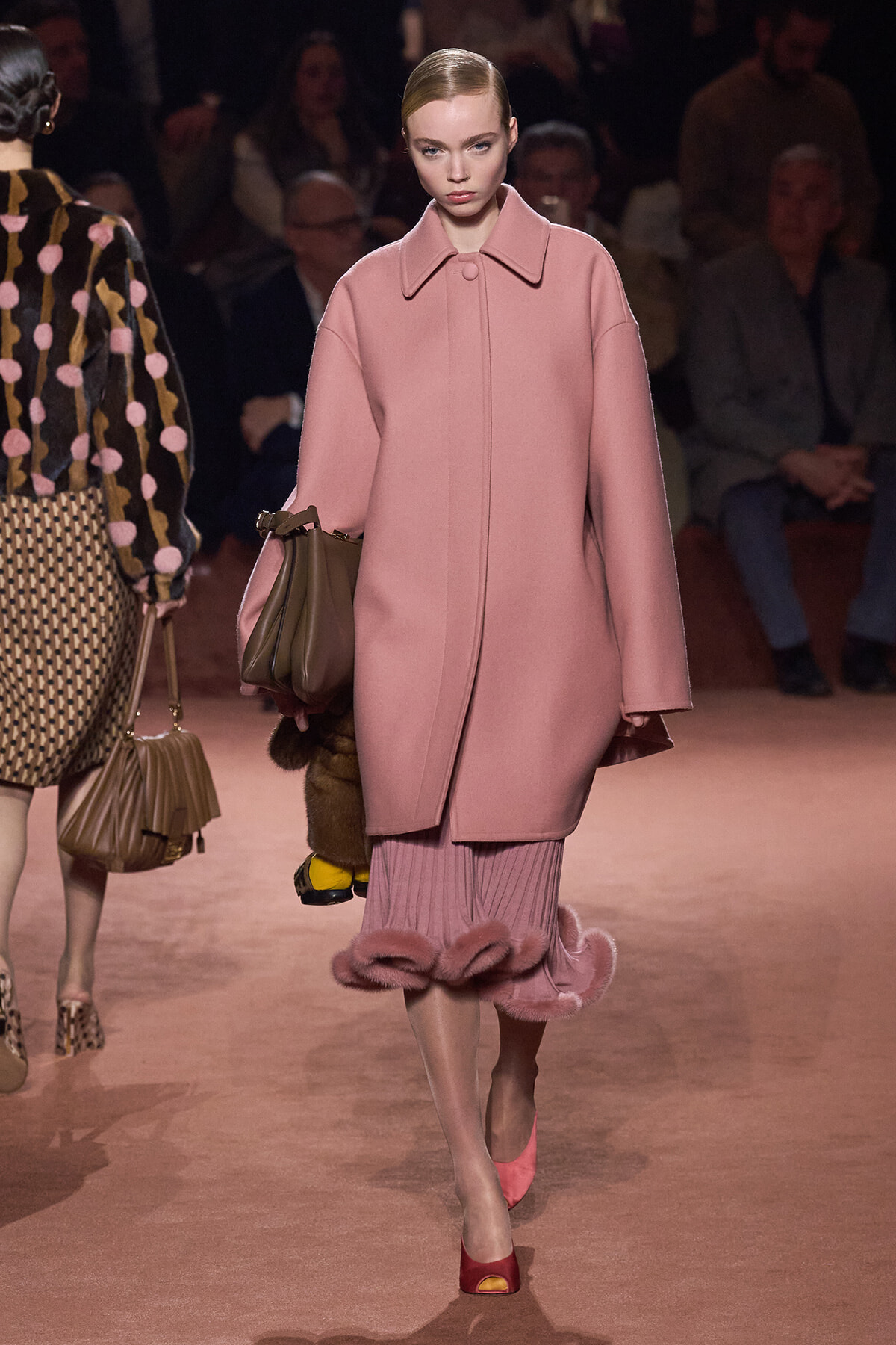 Model walks a runway in a oversized pink coat, carrying a brown handbag, with pink ruffled hem visible beneath.