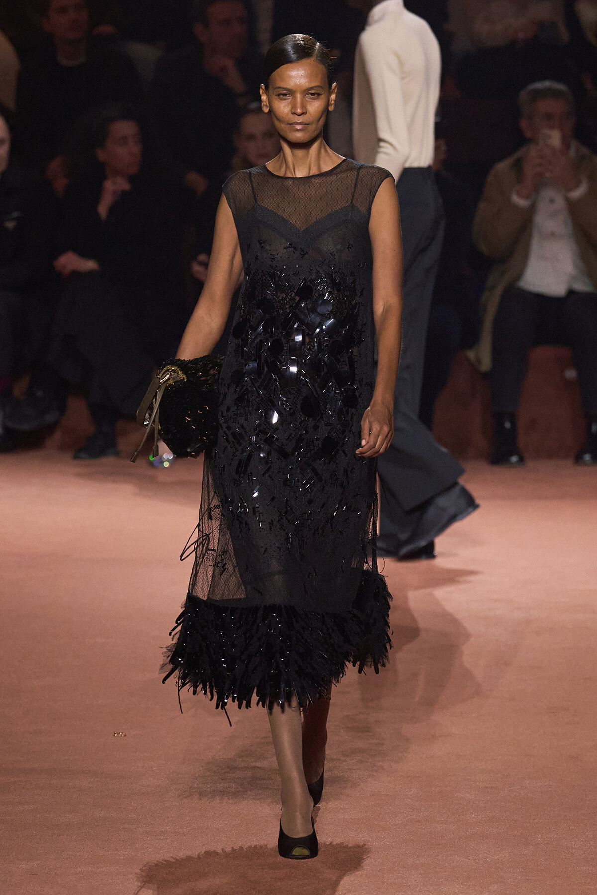 Model walking a runway in a sheer black sequined dress with a feathered hem, carrying a black clutch.