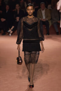 Model walks a fashion show in a black lace dress with sheer sleeves, carrying a small multicolor handbag on a peach runway.