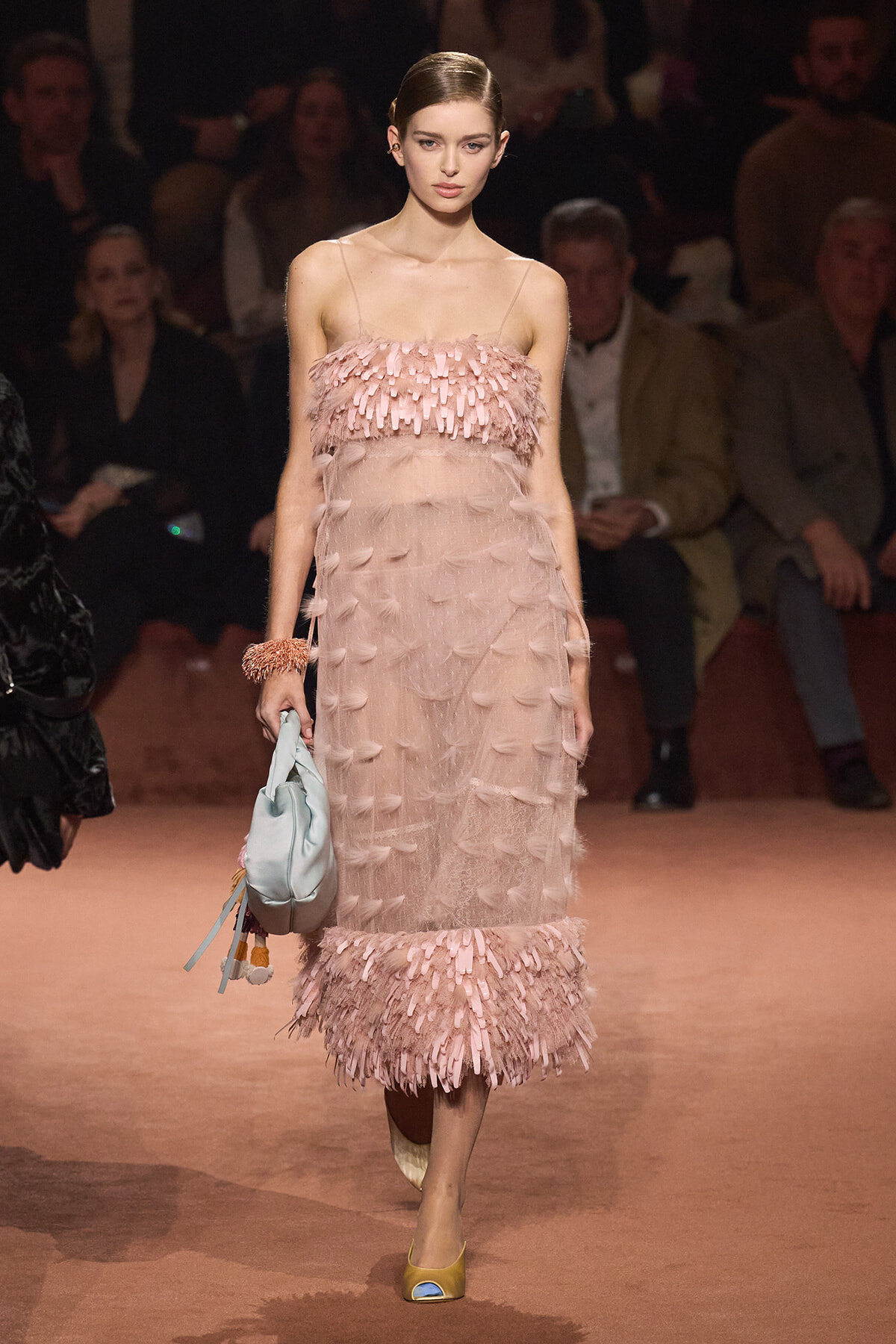 Model on the runway wearing a pale pink fringe dress with a sheer overlay, holding a light blue handbag.