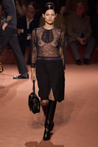 Model walks a fashion runway in a sheer black lace blouse, high-waisted black skirt, and tall black boots, carrying a black patent handbag.