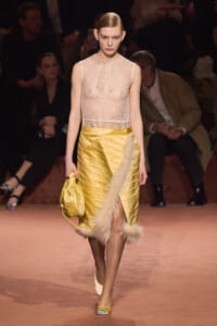 Model walks the runway in a pale lace sleeveless top and a yellow satin banded skirt with fur trim, carrying a gold bag.