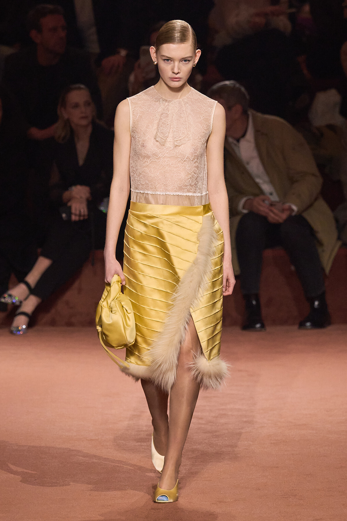 Model walks the runway in a pale lace sleeveless top and a yellow satin banded skirt with fur trim, carrying a gold bag.