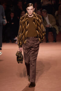Runway model wearing a brown chevron fur coat over a brown sweater, satin brown pants, and carrying a striped brown handbag.