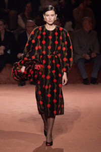 Model walks runway in a black dress with red pom-pom dots and olive-green stripes, belted at the waist, carrying a large matching handbag.