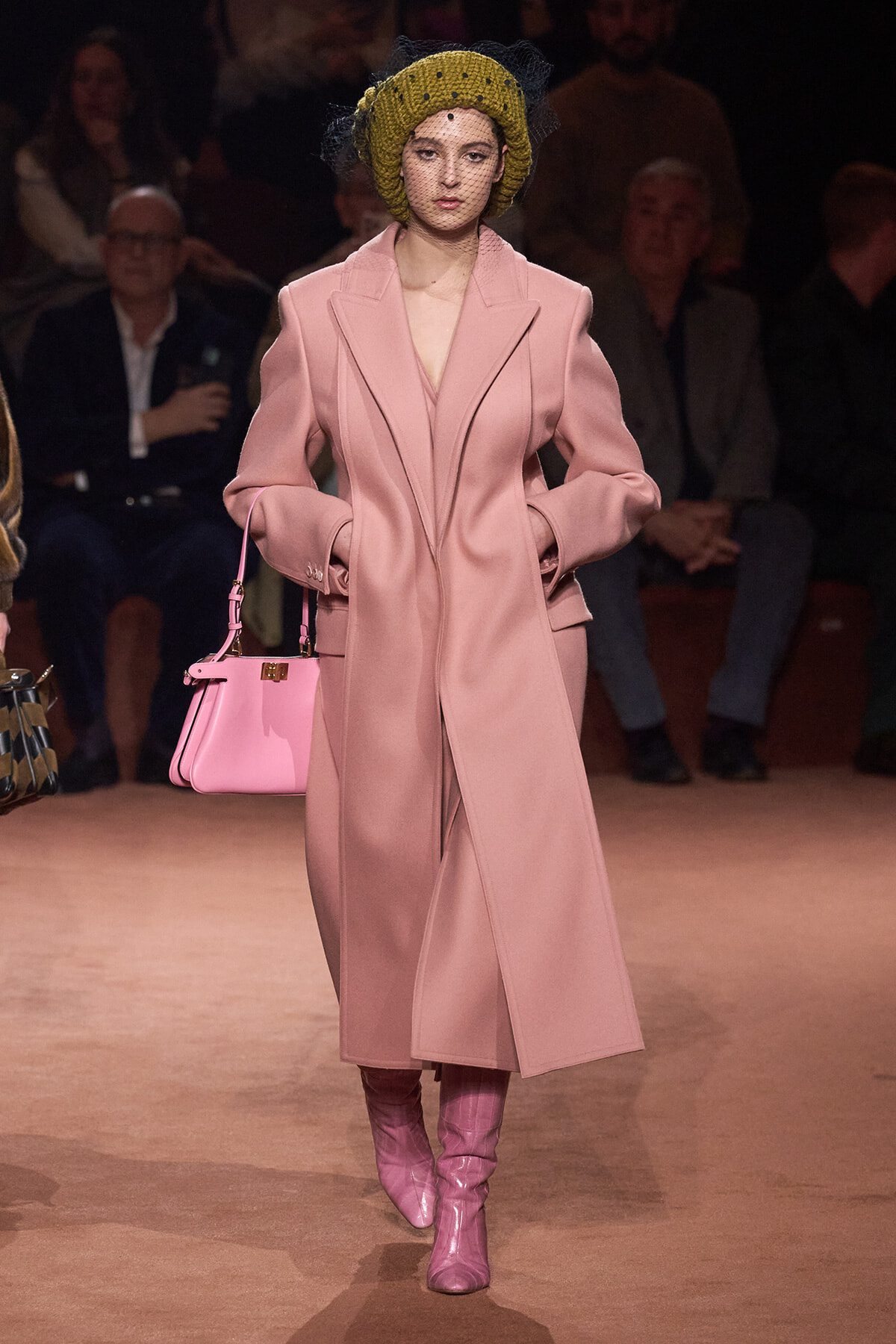 Model walking a fashion runway in a pale pink long coat with matching pink boots and handbag, wearing a yellow knit turban with veil.