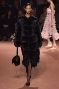 Model on a runway wearing a black fur coat, carrying a dark fur handbag during a fashion show.