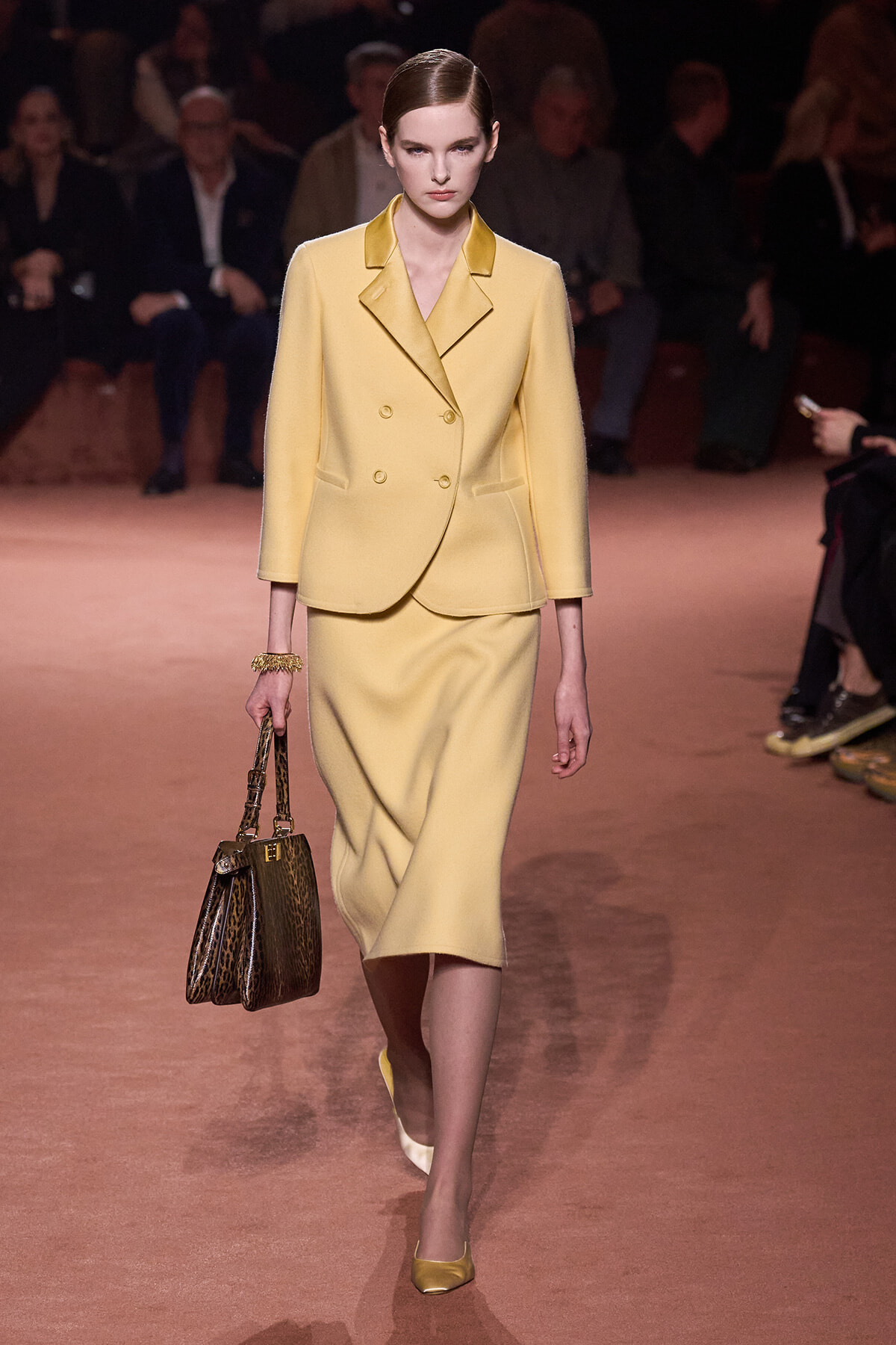 Model walks the runway in a pale yellow double-breasted blazer and matching midi skirt, carrying a leopard print handbag.