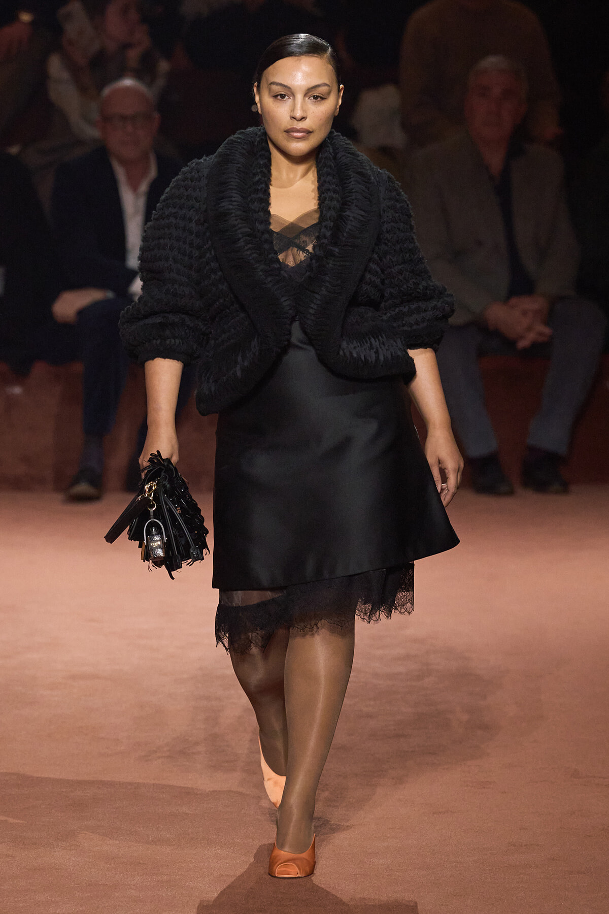 Model walking a runway in a black textured cardigan over a satin skirt with lace hem, tan tights, orange peep-toe heels, carrying a black fringed handbag.