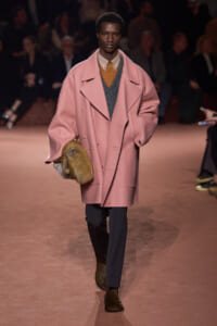 Male model on a runway wearing an oversized pink coat over a gray vest and tan shirt, navy pants, and carrying a fur clutch.