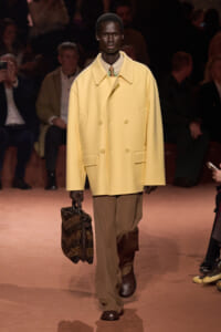 Male model on a runway wearing a oversized pale yellow double-breasted coat, tan wide-leg pants, brown shoes, and carrying a brown textured bag.