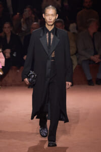 Male model walks a runway in a long black coat over a sheer lace shirt, black trousers, and embellished loafers, holding a small black clutch.