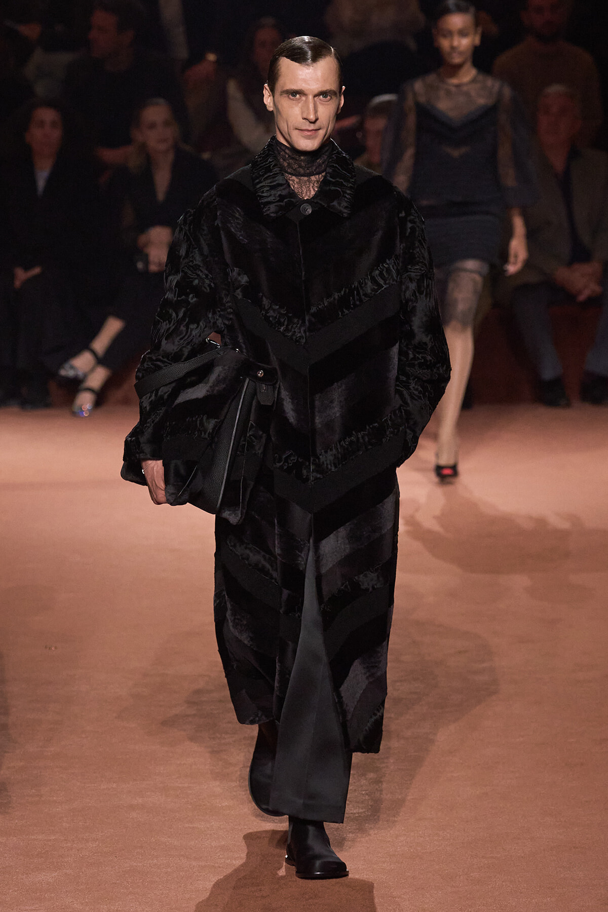Male model on a runway wearing a black fur coat and carrying a black leather bag, audience in the background