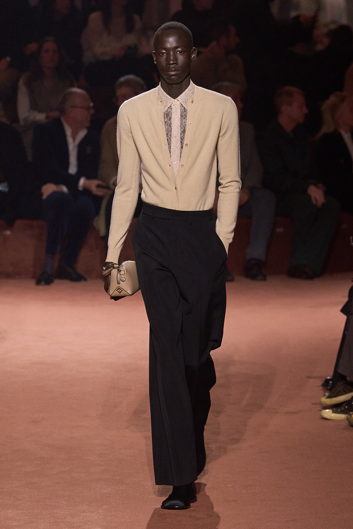 Male model on a runway wearing a beige cardigan over a patterned shirt, black wide-leg trousers, and a beige clutch; audience visible in the background.