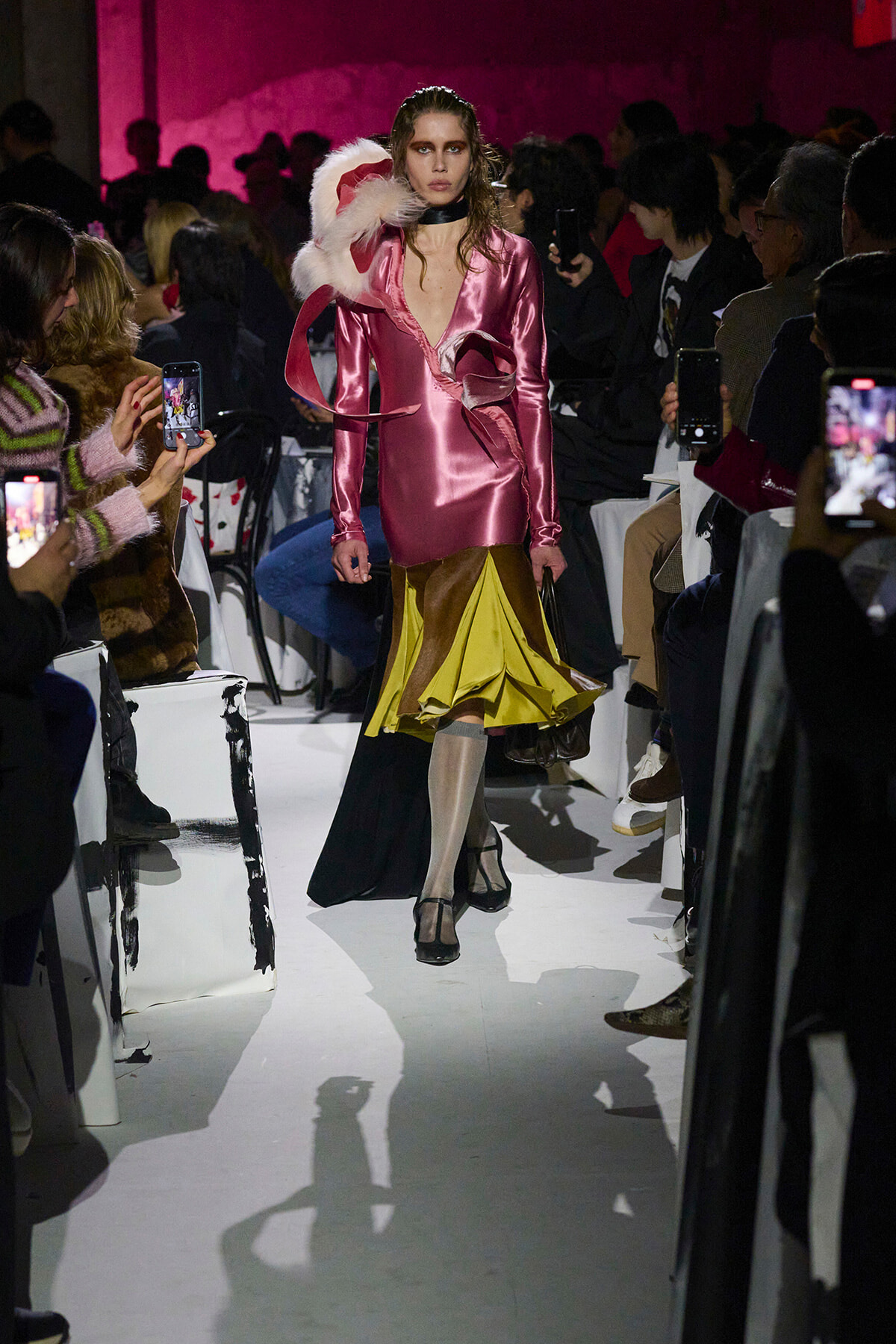 Model walks the runway in a shiny pink metallic dress with a yellow ruffled hem and a large white feather accessory, flanked by spectators taking photos.