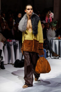 Model walks the runway wearing a two-tone fur stole, mustard knit cardigan, and copper-brown velvet wide-leg pants, carrying a tan leather bag.