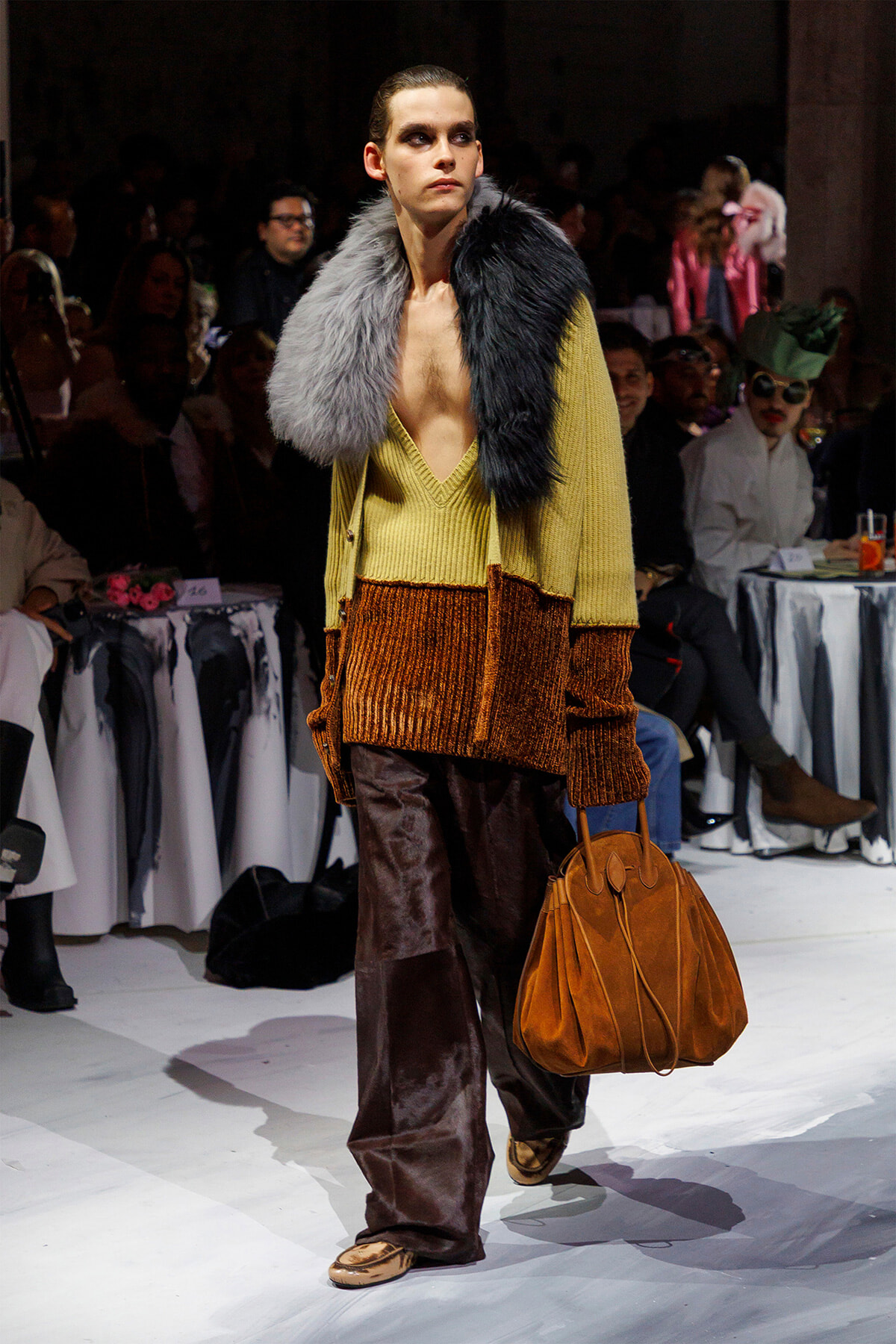 Model walks the runway wearing a two-tone fur stole, mustard knit cardigan, and copper-brown velvet wide-leg pants, carrying a tan leather bag.