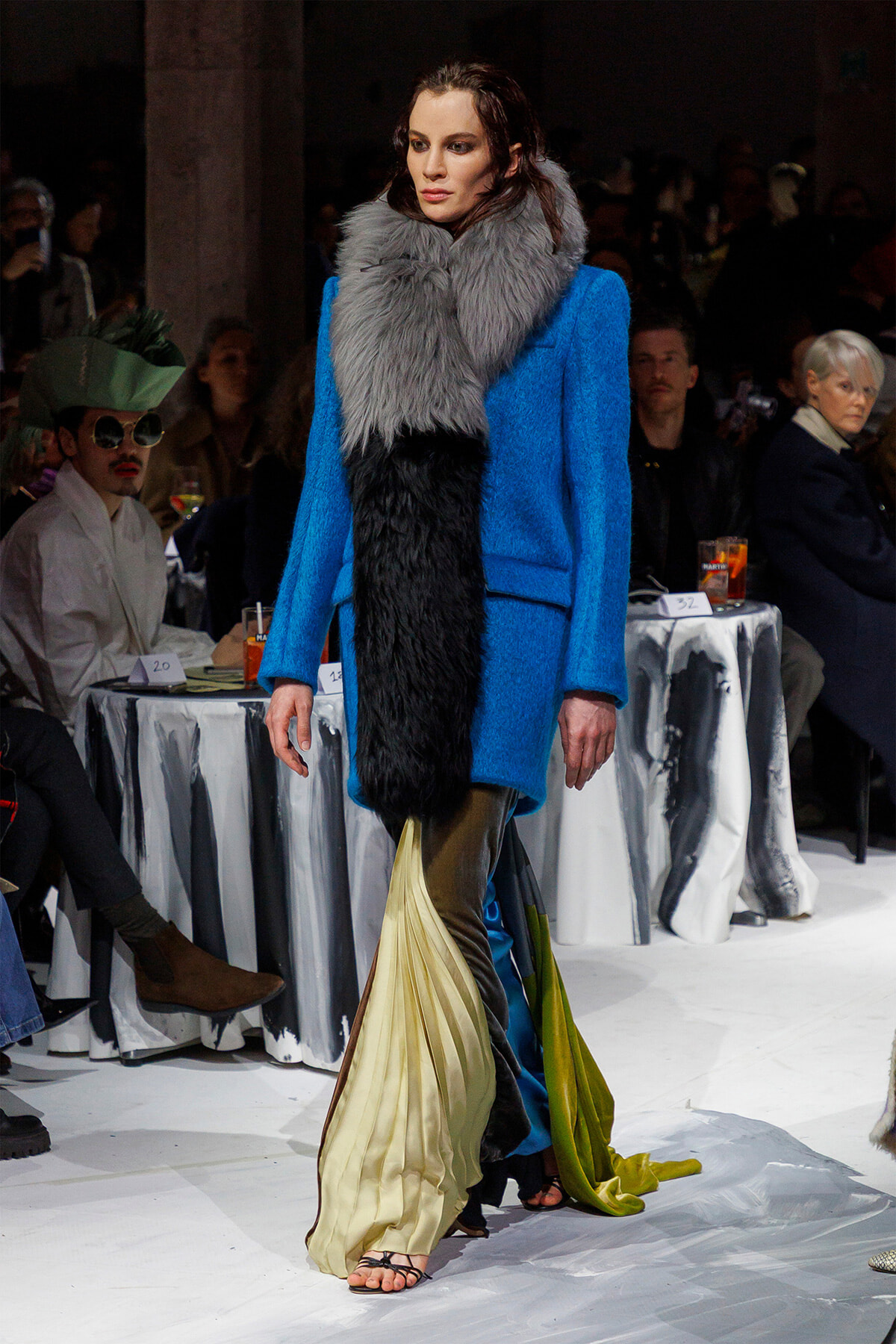 Model on a runway wearing a bright blue wool coat with an oversized gray fur scarf and a flowing gold-olive pleated skirt.
