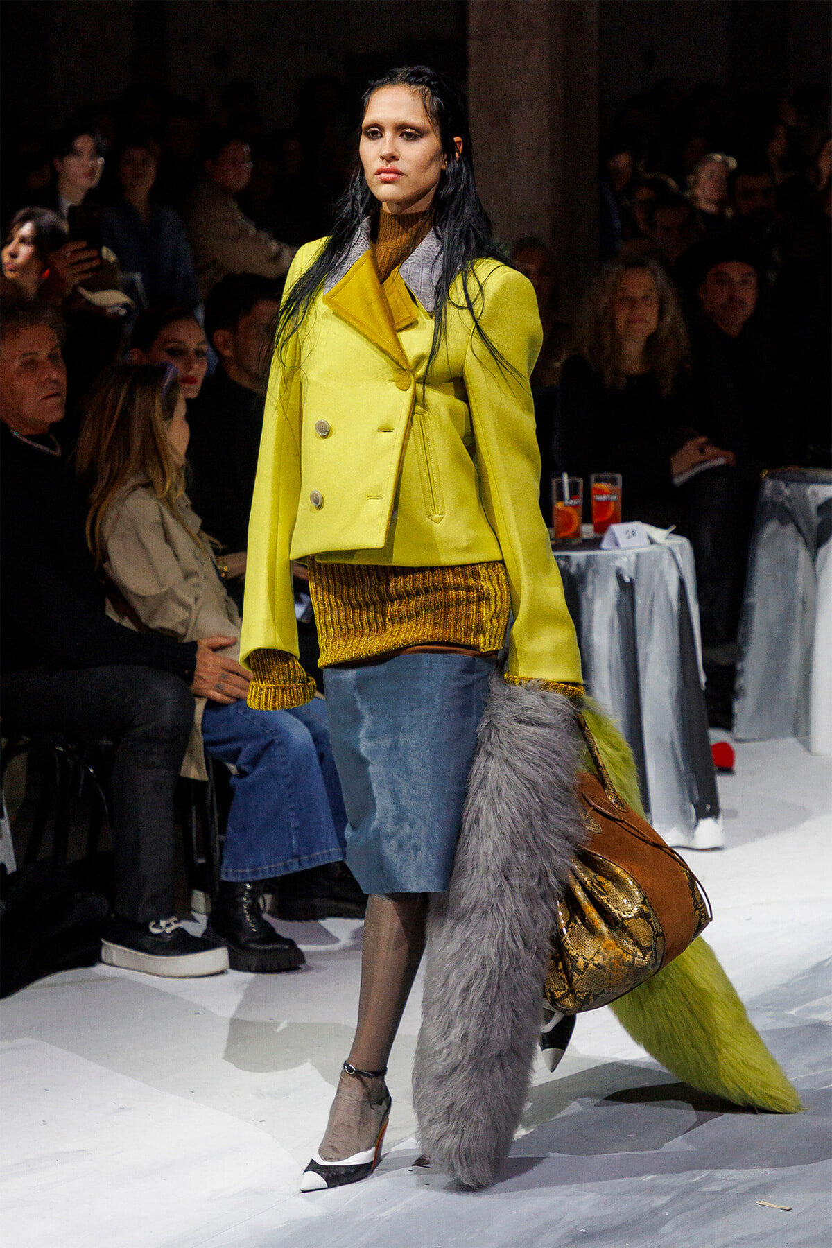 Model walks the runway in a bright yellow double-breasted coat, mustard knit top, denim skirt, and a large colorful handbag with a gray fur scarf trailing behind.