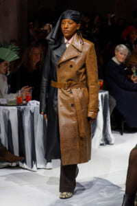Model walking a fashion show in a two-tone trench coat: brown crocodile-embossed side belted at the waist, black leather side, paired with a black head scarf and snakeskin shoes.