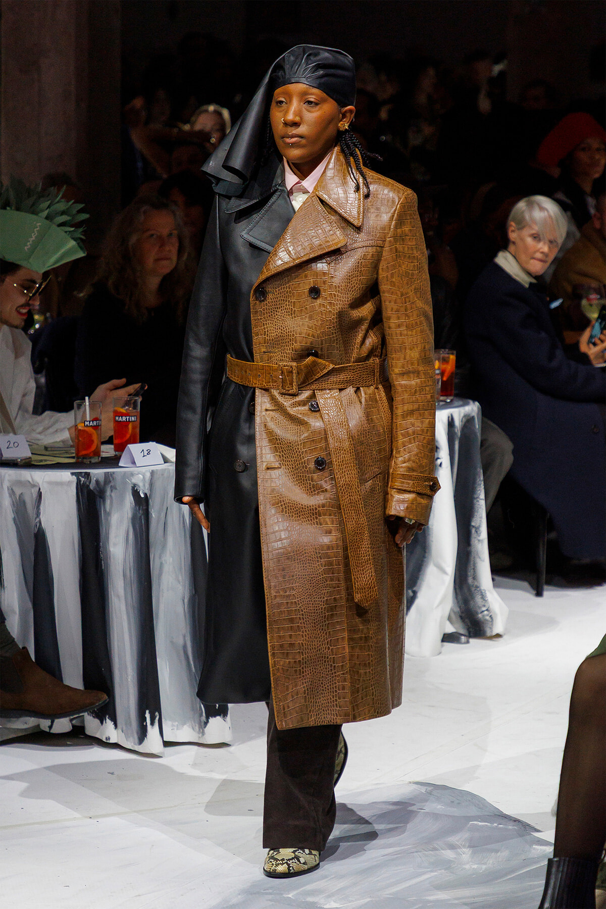 Model walking a fashion show in a two-tone trench coat: brown crocodile-embossed side belted at the waist, black leather side, paired with a black head scarf and snakeskin shoes.