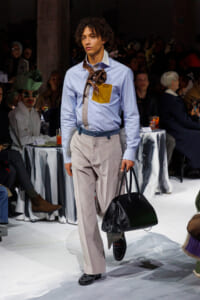 Male model on a runway wearing a light blue shirt, beige trousers, a brown scarf necklace, and a blue belt, carrying a black handbag.