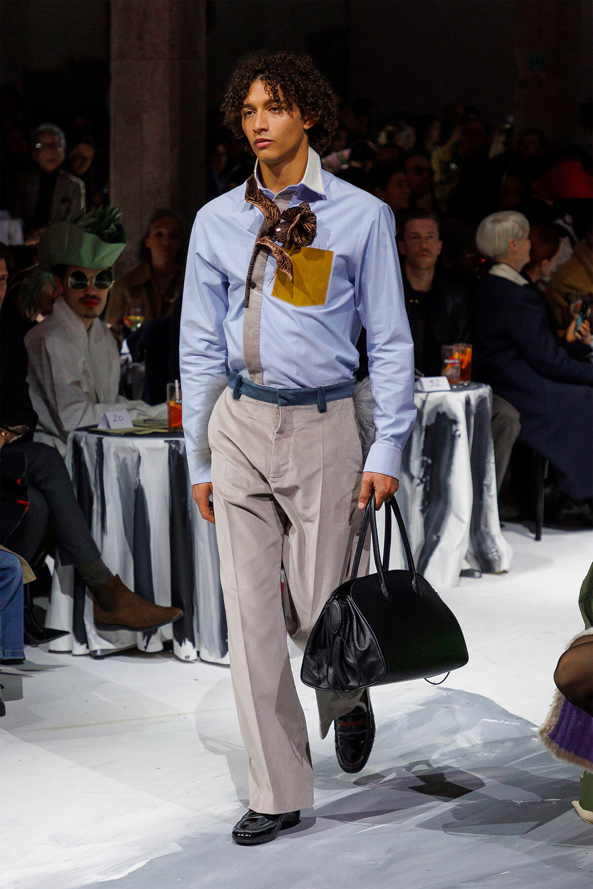 Male model on a runway wearing a light blue shirt, beige trousers, a brown scarf necklace, and a blue belt, carrying a black handbag.
