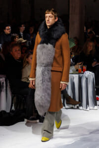 Male model walks a runway in a camel coat with a large two-tone fur scarf (black near the neck, gray down the front).