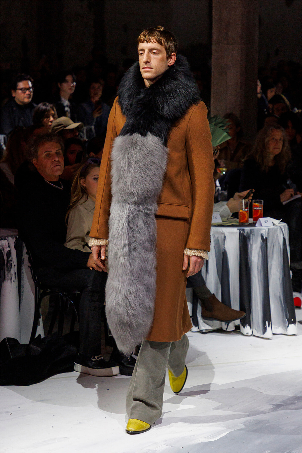 Male model walks a runway in a camel coat with a large two-tone fur scarf (black near the neck, gray down the front).