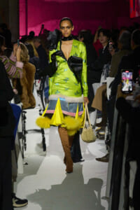 Model walks the runway in a lime-green satin dress with bird prints, black bow details, fur trim, yellow fringe, brown knee-high boots, and a beige handbag.