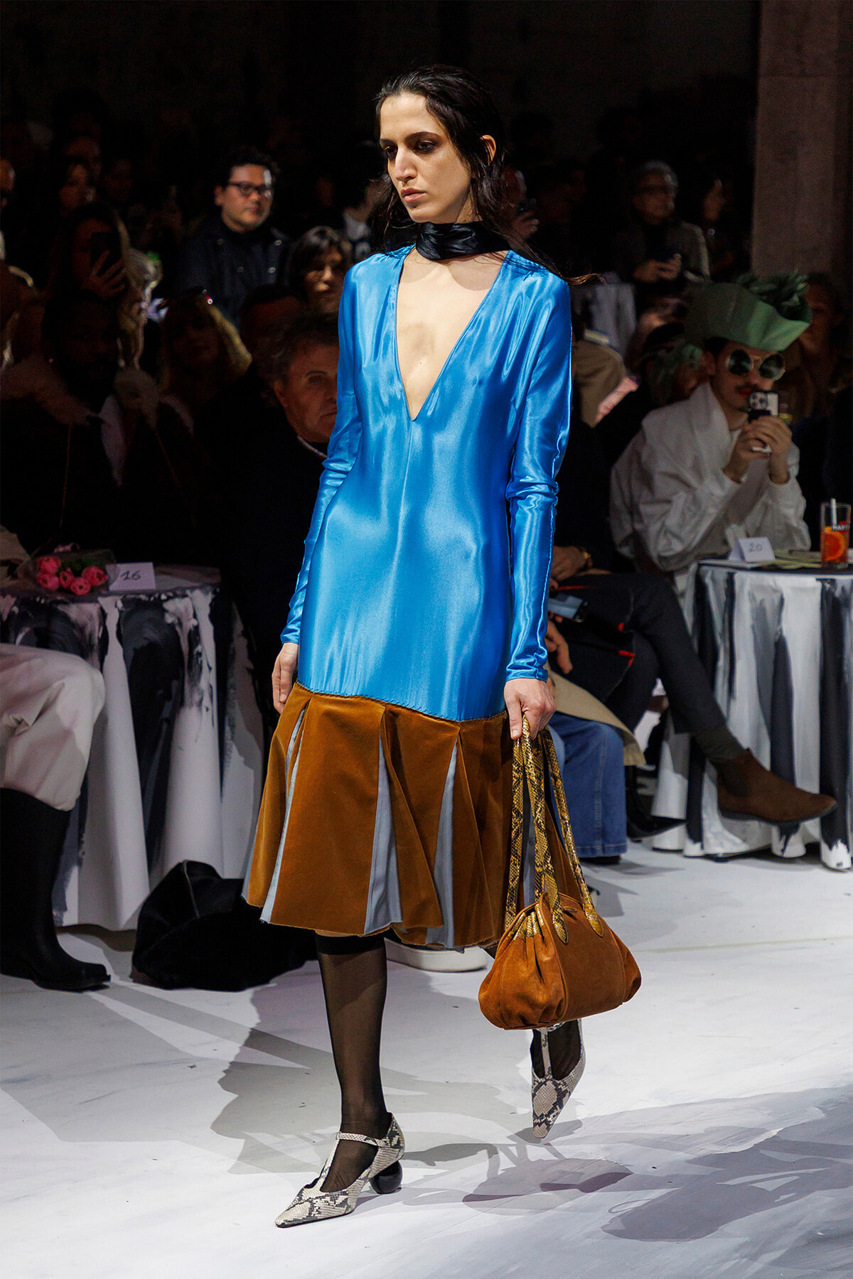 Runway model wearing a cobalt blue satin deep-V dress with a brown pleated skirt, black tights, snakeskin heels, and a brown handbag.