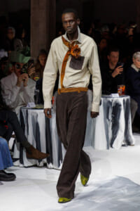 Male model walking a fashion runway in a cream shirt with an orange neck scarf and brown wide-leg pants, accented by a decorative orange flower at the chest pocket.