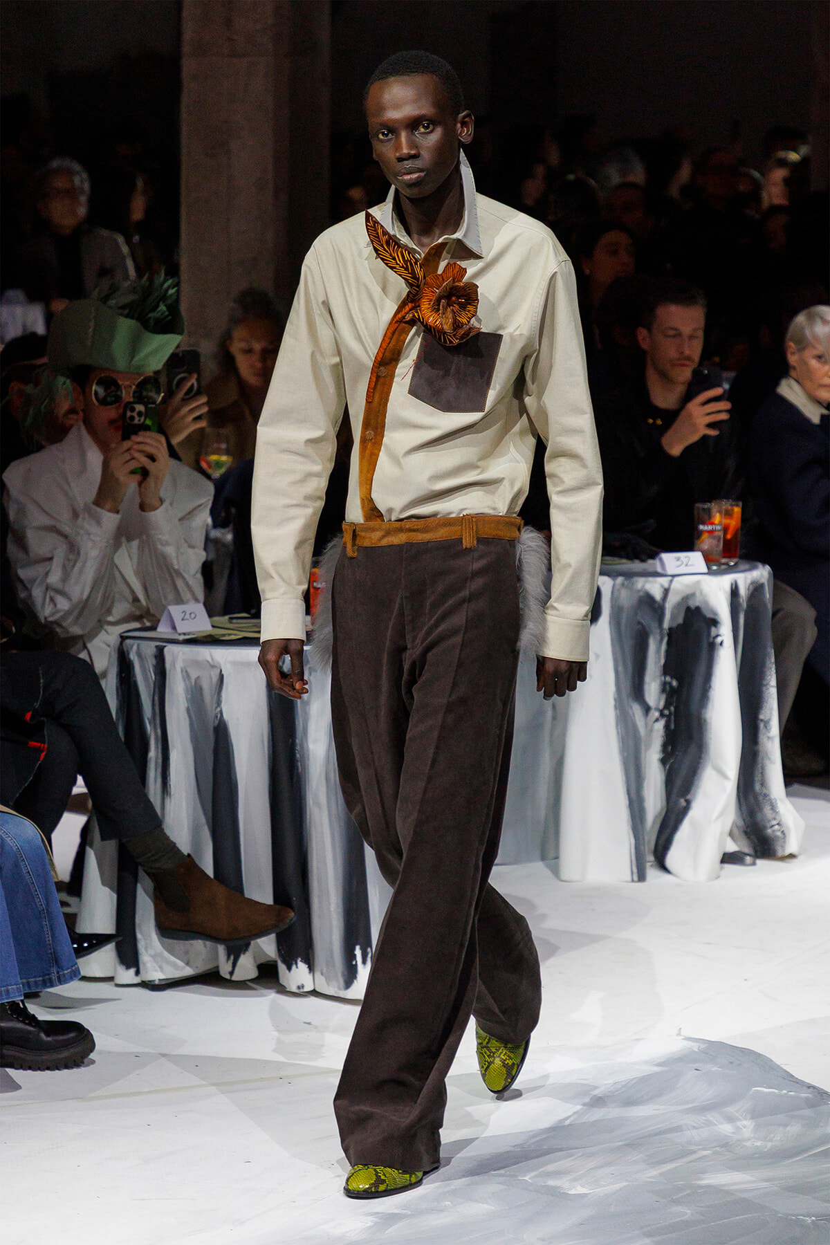 Male model walking a fashion runway in a cream shirt with an orange neck scarf and brown wide-leg pants, accented by a decorative orange flower at the chest pocket.