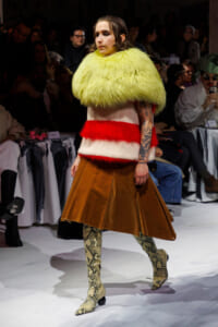 Model on runway wearing a neon yellow fur cape over a red-and-white knit top and a brown A-line leather skirt, with snakeskin boots; audience in background.