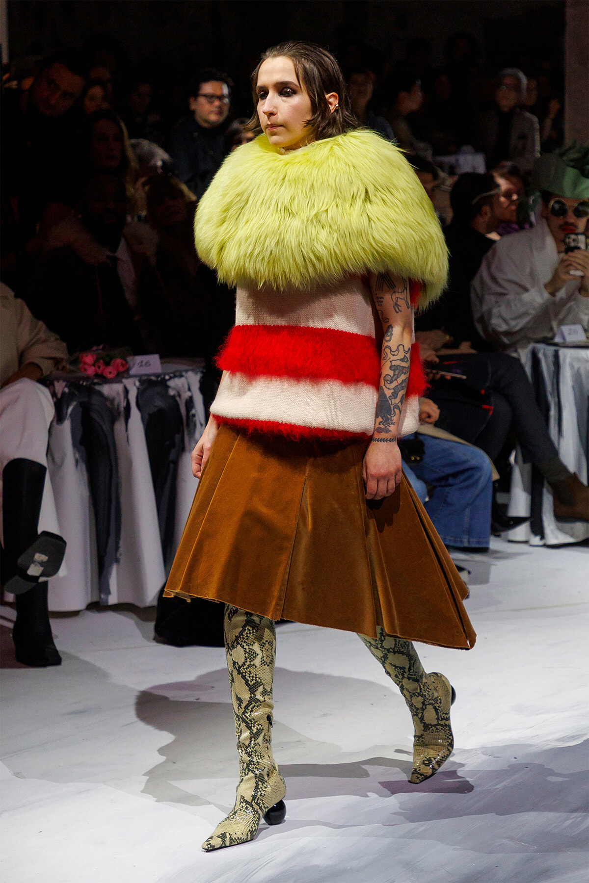 Model on runway wearing a neon yellow fur cape over a red-and-white knit top and a brown A-line leather skirt, with snakeskin boots; audience in background.