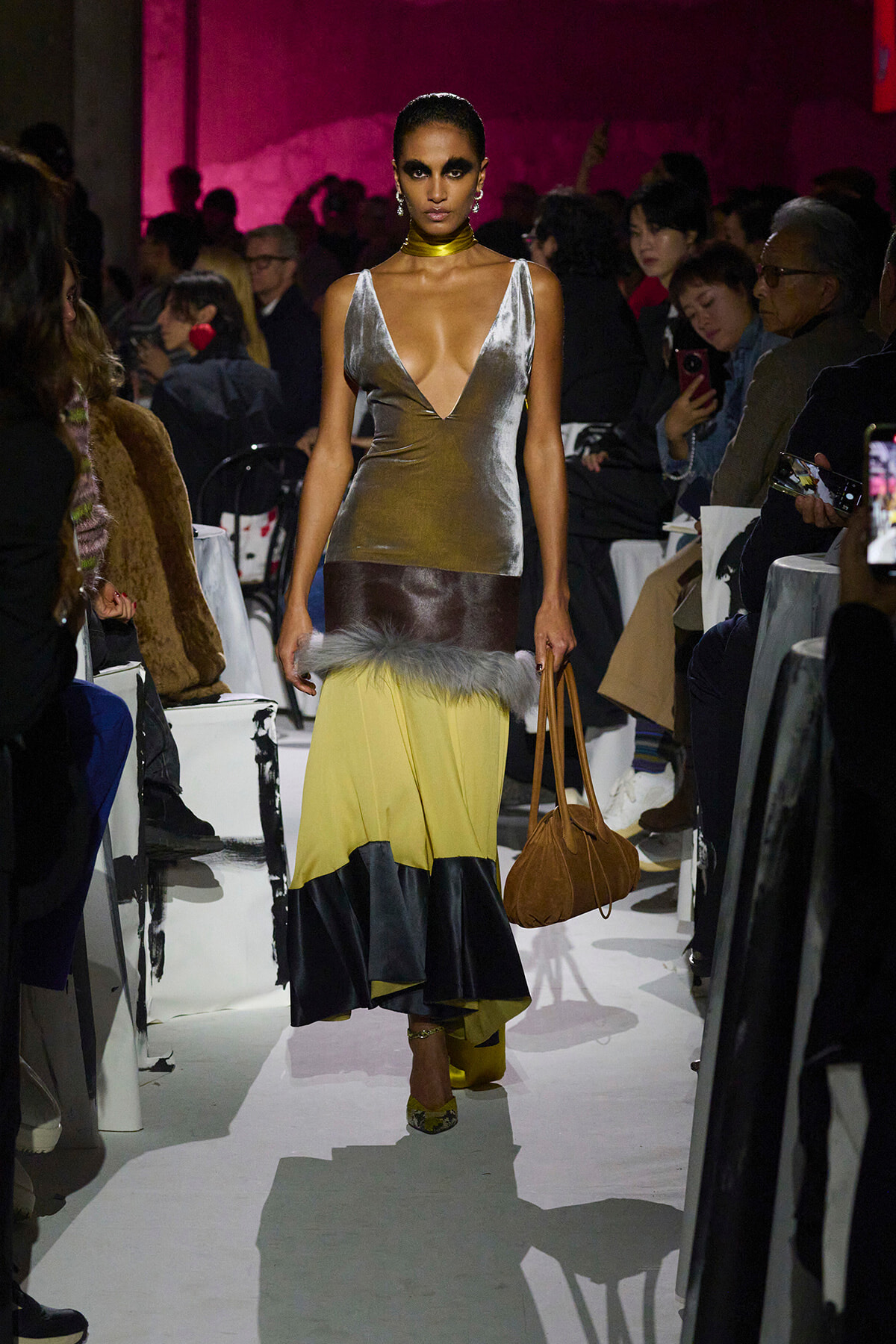 Model walks a runway wearing a metallic silver plunging top and a color-block yellow-to-black skirt with gray fur trim, carrying a tan handbag.