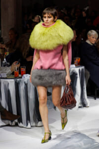 Model walking a fashion runway in a pink dress with a large neon yellow fur collar and gray fur hem, carrying a brown bag.