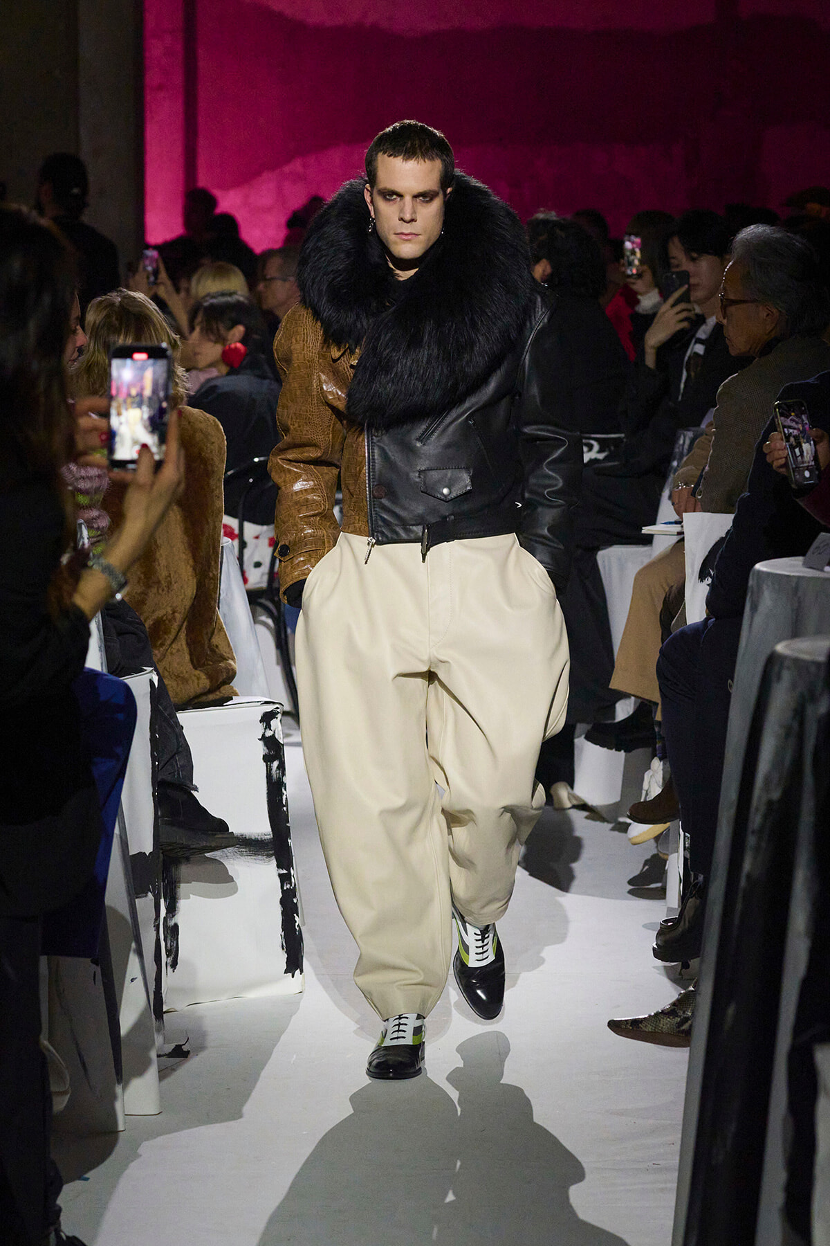 Male model on a runway wearing a black fur-curled leather jacket with beige/cream wide trousers, spectators filming.